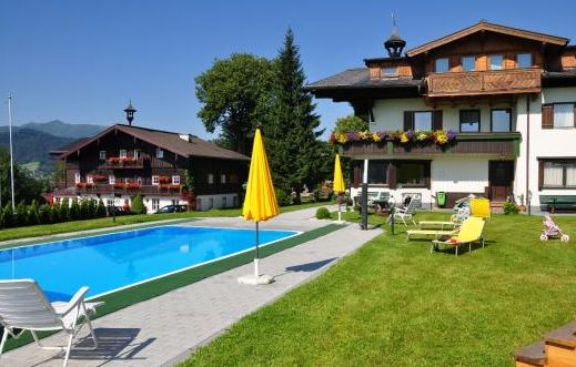 Außenpool mit gelben Sonnenschirmen, Liegestühlen und zwei Gebäuden im alpinen Stil, umgeben von grünem Gras und Bäumen unter einem klaren blauen Himmel.