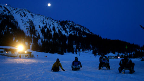 Vier Personen rodeln nachts bei Vollmond auf einem schneebedeckten Hang, im Hintergrund sind Berge und ein helles Licht zu sehen.