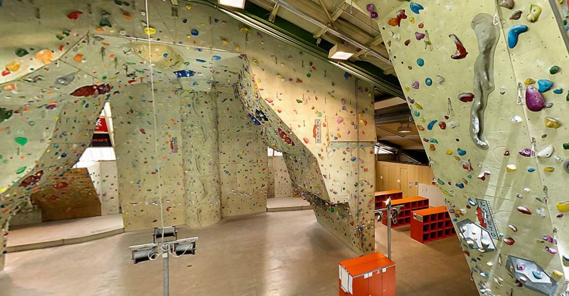 Indoor-Kletterhalle mit hohen Kletterwänden mit bunten Griffen, einer Glasbarriere, Schließfächern und Kletterausrüstung auf einem polierten Betonboden.