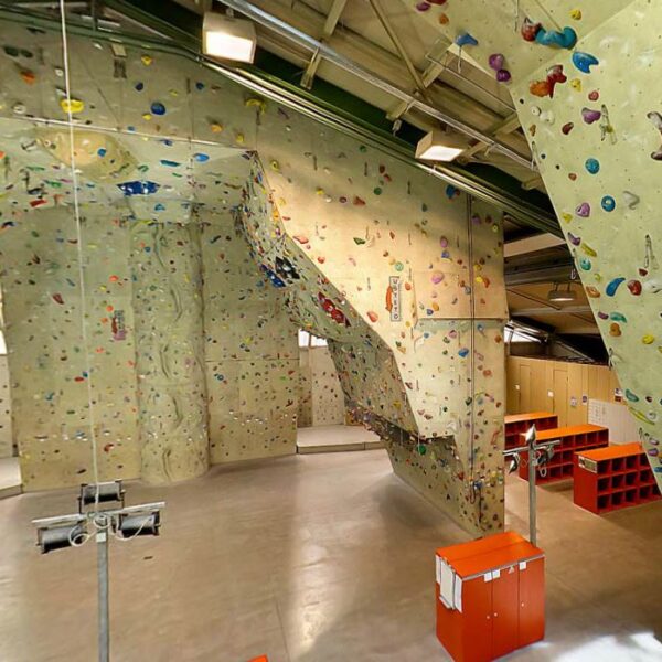 Indoor-Kletterhalle mit hohen Kletterwänden mit bunten Griffen, einer Glasbarriere, Schließfächern und Kletterausrüstung auf einem polierten Betonboden.