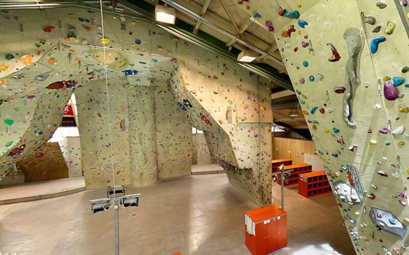 Indoor-Kletterhalle mit hohen Kletterwänden mit bunten Griffen, einer Glasbarriere, Schließfächern und Kletterausrüstung auf einem polierten Betonboden.