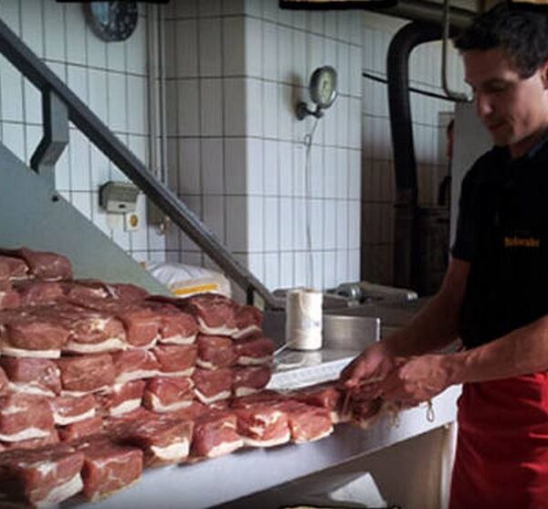 Ein Mann in einem schwarzen Hemd und einer roten Schürze bereitet Stapel rohen Fleisches vor und arrangiert sie auf einem Tisch in einem gefliesten Raum, möglicherweise einer Metzgerei oder einem Fleischverarbeitungsbetrieb.