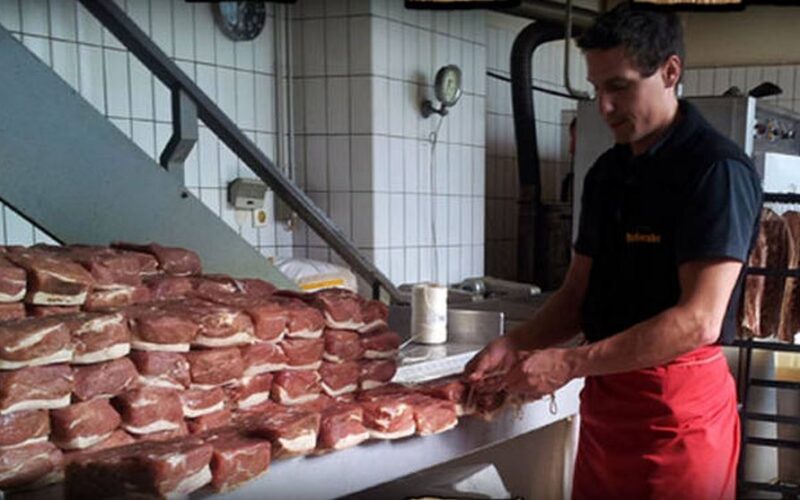 Ein Mann in einem schwarzen Hemd und einer roten Schürze bereitet Stapel rohen Fleisches vor und arrangiert sie auf einem Tisch in einem gefliesten Raum, möglicherweise einer Metzgerei oder einem Fleischverarbeitungsbetrieb.