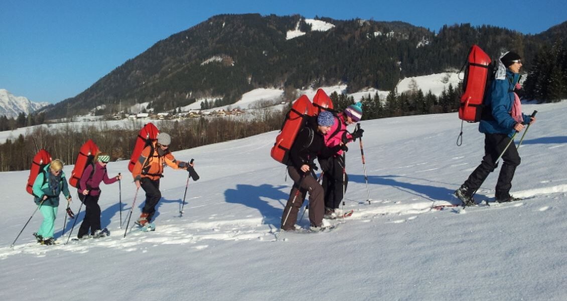 Eine Gruppe von sechs Personen läuft mit Schneeschuhen in einer verschneiten Landschaft bergauf. Sie tragen Rucksäcke und Winterkleidung, im Hintergrund sind Berge und Bäume zu sehen.