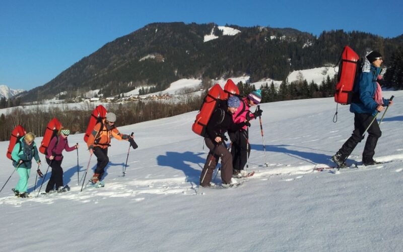 Eine Gruppe von sechs Personen läuft mit Schneeschuhen in einer verschneiten Landschaft bergauf. Sie tragen Rucksäcke und Winterkleidung, im Hintergrund sind Berge und Bäume zu sehen.