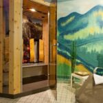 Eine kleine Saunakabine mit Glastür steht neben einem Liegestuhl und einem Beistelltisch, an der Wand ein gemaltes Gebirgswandbild und in der Nähe gestapelte Felsen.