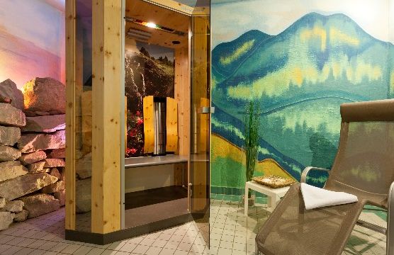 Eine kleine Saunakabine mit Glastür steht neben einem Liegestuhl und einem Beistelltisch, an der Wand ein gemaltes Gebirgswandbild und in der Nähe gestapelte Felsen.