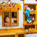 Ein kleines Kind schaut aus einem Fenster in einem Spielhaus, das mit bunten Bildern von Bauernhoftieren und Heu geschmückt ist.