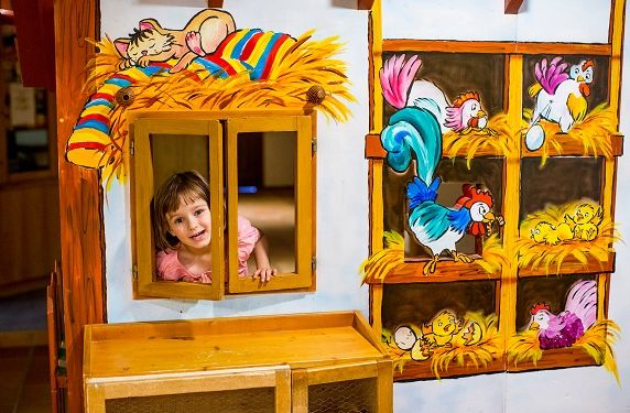 Ein kleines Kind schaut aus einem Fenster in einem Spielhaus, das mit bunten Bildern von Bauernhoftieren und Heu geschmückt ist.