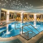 Hallenbadbereich mit Liegestühlen, sternförmiger Deckenbeleuchtung, großen Fenstern und einem kleinen Wasserfall auf der linken Seite.