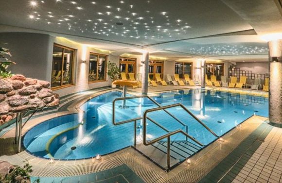 Hallenbadbereich mit Liegestühlen, sternförmiger Deckenbeleuchtung, großen Fenstern und einem kleinen Wasserfall auf der linken Seite.