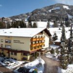 Hotelgebäude mit schneebedecktem Dach und Umgebung, geparkte Autos, Kiefern und Berge im Hintergrund an einem klaren Wintertag.