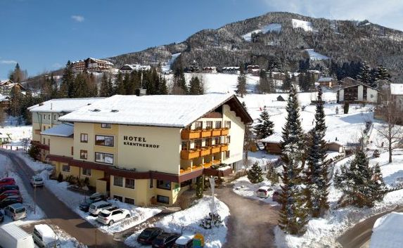 Hotelgebäude mit schneebedecktem Dach und Umgebung, geparkte Autos, Kiefern und Berge im Hintergrund an einem klaren Wintertag.