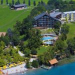 Luftaufnahme eines Seeufer-Resorts mit mehreren Gebäuden, Außenpools, Liegestühlen und Sonnenschirmen, umgeben von Bäumen und Grasflächen neben dem klaren blauen Wasser.