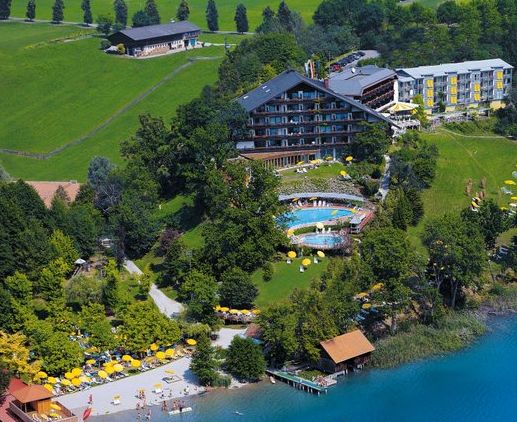 Luftaufnahme eines Seeufer-Resorts mit mehreren Gebäuden, Außenpools, Liegestühlen und Sonnenschirmen, umgeben von Bäumen und Grasflächen neben dem klaren blauen Wasser.
