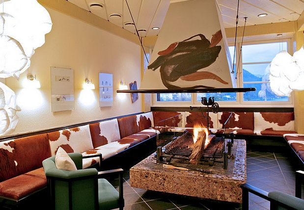 Eine gemütliche Lounge mit mit Kuhfell gepolsterten Bänken, einem zentralen offenen Kamin, abstrakter Kunst und großen Fenstern mit Blick auf die Berge.