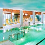 Hallenbad mit klarem Wasser, Liegestühlen und großen Fenstern, die natürliches Licht hereinlassen; weiße Säulen und eine Holzdecke sind sichtbar.