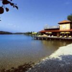 Ein Gebäude am See mit einer Terrasse, gelben Sonnenschirmen und Liegestühlen liegt an einem klaren, ruhigen Wasser und einem Kiesstrand unter blauem Himmel.