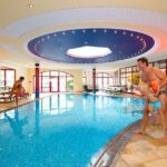 Ein Erwachsener und zwei Kinder spielen an einem Hallenbad mit Liegestühlen und großen Fenstern, die den Bereich umgeben.