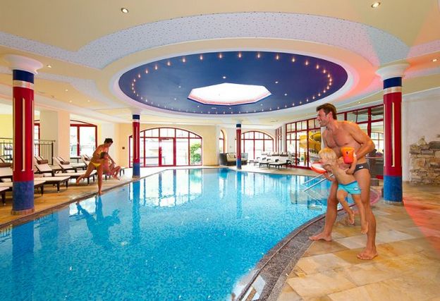 Ein Erwachsener und zwei Kinder spielen an einem Hallenbad mit Liegestühlen und großen Fenstern, die den Bereich umgeben.