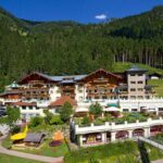 Großer alpiner Hotelkomplex mit mehreren Balkonen und Terrassen, umgeben von bewaldeten Bergen, einem Parkplatz und einer gepflegten Anlage unter blauem Himmel.