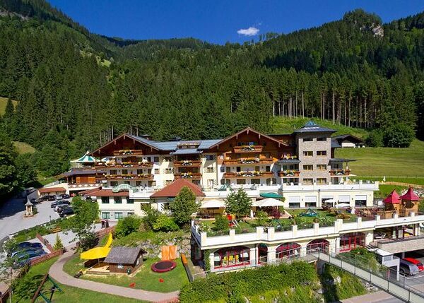 Großer alpiner Hotelkomplex mit mehreren Balkonen und Terrassen, umgeben von bewaldeten Bergen, einem Parkplatz und einer gepflegten Anlage unter blauem Himmel.