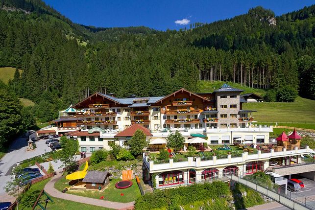 Großer alpiner Hotelkomplex mit mehreren Balkonen und Terrassen, umgeben von bewaldeten Bergen, einem Parkplatz und einer gepflegten Anlage unter blauem Himmel.