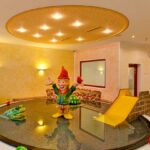 Indoor-Kinderspielbereich mit einem flachen, runden Becken, einer Rutsche, buntem Spielzeug und einer großen Gnom-Statue in der Mitte unter einer runden Deckenleuchte.