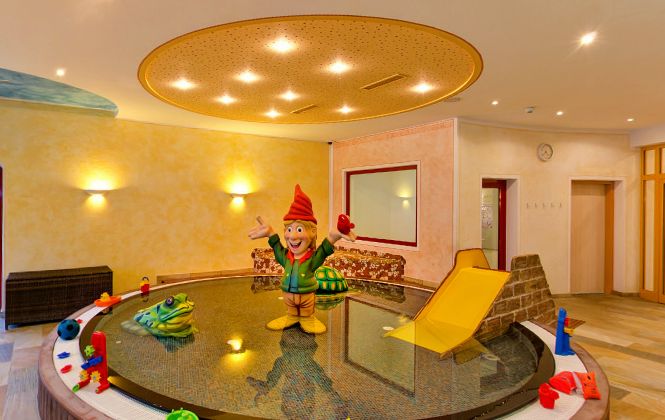 Indoor-Kinderspielbereich mit einem flachen, runden Becken, einer Rutsche, buntem Spielzeug und einer großen Gnom-Statue in der Mitte unter einer runden Deckenleuchte.
