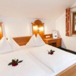 Ein ordentlich gemachtes Hotelbett mit zwei weißen Bettdecken, die mit roten Rosen verziert sind, ein hölzernes Kopfteil, Nachttischlampen, ein Fenster, ein Schreibtisch, ein Heizkörper und gemusterte rote Vorhänge.