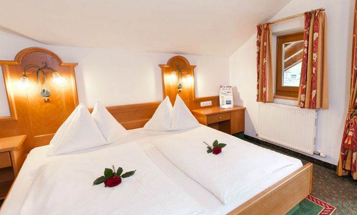 Ein ordentlich gemachtes Hotelbett mit zwei weißen Bettdecken, die mit roten Rosen verziert sind, ein hölzernes Kopfteil, Nachttischlampen, ein Fenster, ein Schreibtisch, ein Heizkörper und gemusterte rote Vorhänge.