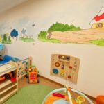 Buntes Kinderspielzimmer mit Spielzeug, einer Wandtafel und einem Wandgemälde mit Bäumen, Tieren und einem Haus mit rotem Dach.