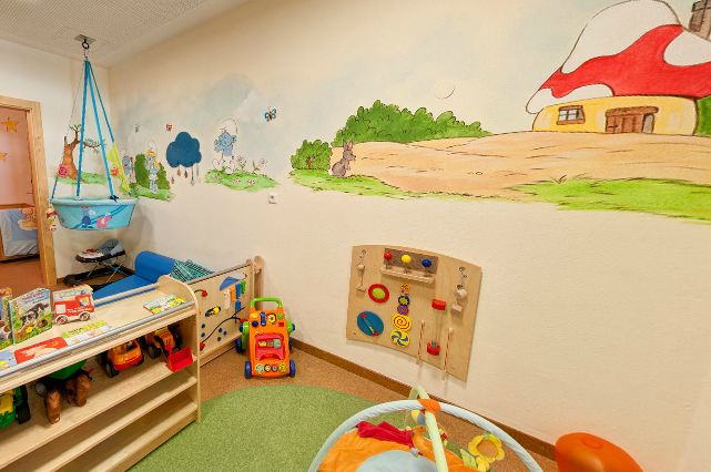 Buntes Kinderspielzimmer mit Spielzeug, einer Wandtafel und einem Wandgemälde mit Bäumen, Tieren und einem Haus mit rotem Dach.