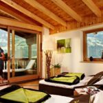 Eine Frau steht auf einem Balkon mit Blick auf die Berge und liest. Drinnen stehen zwei Sessel mit grünen Kissen vor großen Fenstern in einem holzgetäfelten Raum.