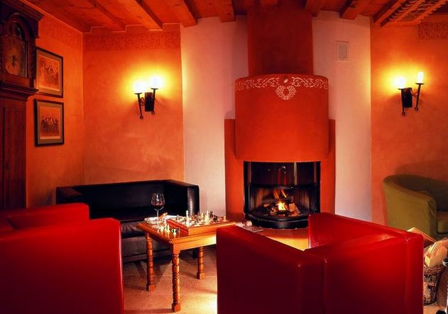 Ein gemütliches Zimmer mit roten Sesseln, einem schwarzen Sofa, einem Holztisch mit Gläsern und einem Schachbrett und einem beleuchteten Kamin in einem orangefarbenen Schornstein.