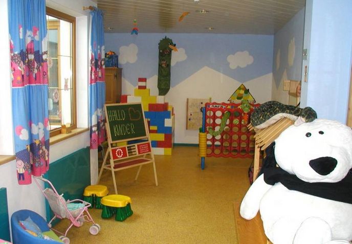 Ein Kinderspielzimmer mit buntem Spielzeug, einer Kreidetafel mit der Aufschrift "HALLO KINDER", einem großen weißen Stoffbären und blauen Vorhängen mit Cartoon-Motiven.
