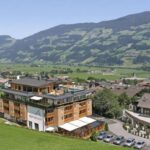 Ein mehrstöckiges Hotelgebäude steht neben einer grünen Wiese mit Bergen und einem Dorf im Hintergrund unter einem klaren blauen Himmel.