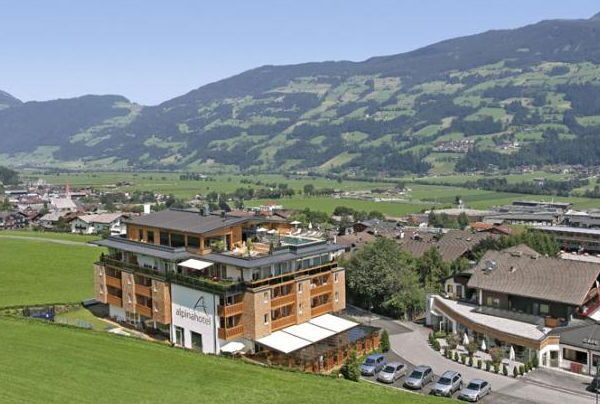 Ein mehrstöckiges Hotelgebäude steht neben einer grünen Wiese mit Bergen und einem Dorf im Hintergrund unter einem klaren blauen Himmel.