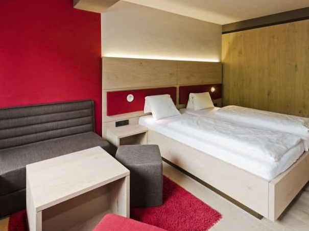 Modernes Hotelzimmer mit einem Doppelbett, zwei weißen Kissen, einem grauen Sofa, einem kleinen Tisch, einer roten Akzentwand und einem Teppich.