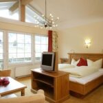 Ein gemütliches Hotelzimmer verfügt über ein Doppelbett, Holzmöbel, einen Fernseher auf einem Ständer, eine kleine Sitzecke, große Fenster und einen modernen Kronleuchter.