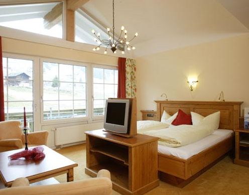 Ein gemütliches Hotelzimmer verfügt über ein Doppelbett, Holzmöbel, einen Fernseher auf einem Ständer, eine kleine Sitzecke, große Fenster und einen modernen Kronleuchter.