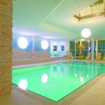 Hallenbad mit grün beleuchtetem Wasser, umgeben von Liegestühlen, Steinsäulen, großen Fenstern und modernem Dekor. Sphärische Deckenleuchten und Pflanzen schmücken den Raum.