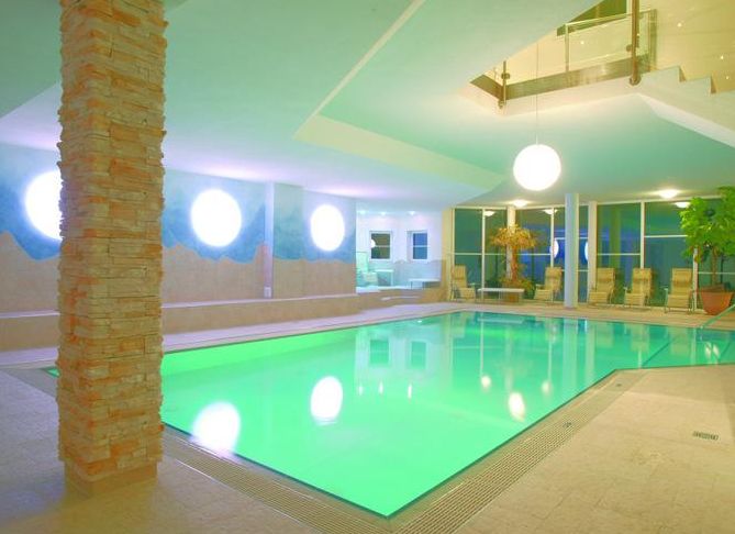 Hallenbad mit grün beleuchtetem Wasser, umgeben von Liegestühlen, Steinsäulen, großen Fenstern und modernem Dekor. Sphärische Deckenleuchten und Pflanzen schmücken den Raum.