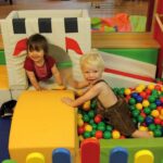 Zwei kleine Kinder spielen in einer mit bunten Plastikbällen gefüllten Ballgrube, umgeben von weichen Spielgeräten.