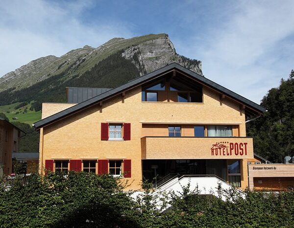 Ein dreistöckiges Gebäude mit der Aufschrift "Hotel Post" und roten Fensterläden vor einer Kulisse aus grünen Bergen und einem teilweise bewölkten Himmel.