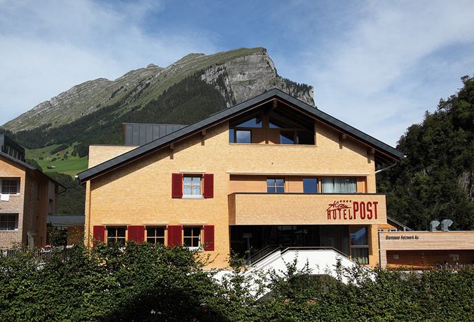 Ein dreistöckiges Gebäude mit der Aufschrift "Hotel Post" und roten Fensterläden vor einer Kulisse aus grünen Bergen und einem teilweise bewölkten Himmel.