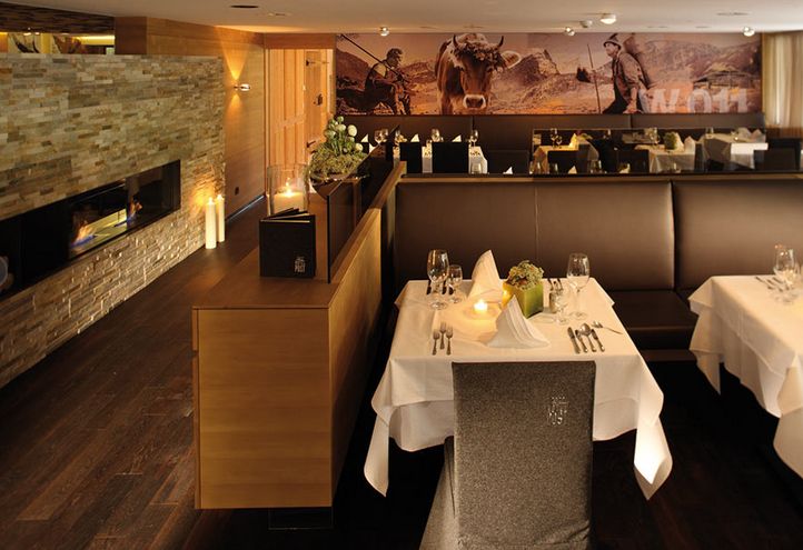 Modernes Restaurantinterieur mit ordentlich gedeckten Tischen, braunen Ledersesseln, einer Akzentwand aus Stein mit Kamin und einem Wandgemälde mit Außenaufnahmen an der Rückwand.