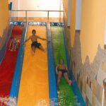 Drei Personen rutschen auf parallelen Wasserrutschen in den Farben Rot, Gelb und Grün hinunter, umgeben von hellbraunen Kachelwänden und Säulen.