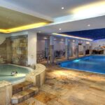 Wellnessbereich im Innenbereich mit einem runden Whirlpool und einem rechteckigen Schwimmbecken, mit Steinfliesenboden und einer Decke mit eingebauter Beleuchtung.