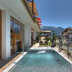 Außenpool neben einem modernen Gebäude mit Liegestühlen und roten Sonnenschirmen vor der Kulisse der Berge und des klaren blauen Himmels.